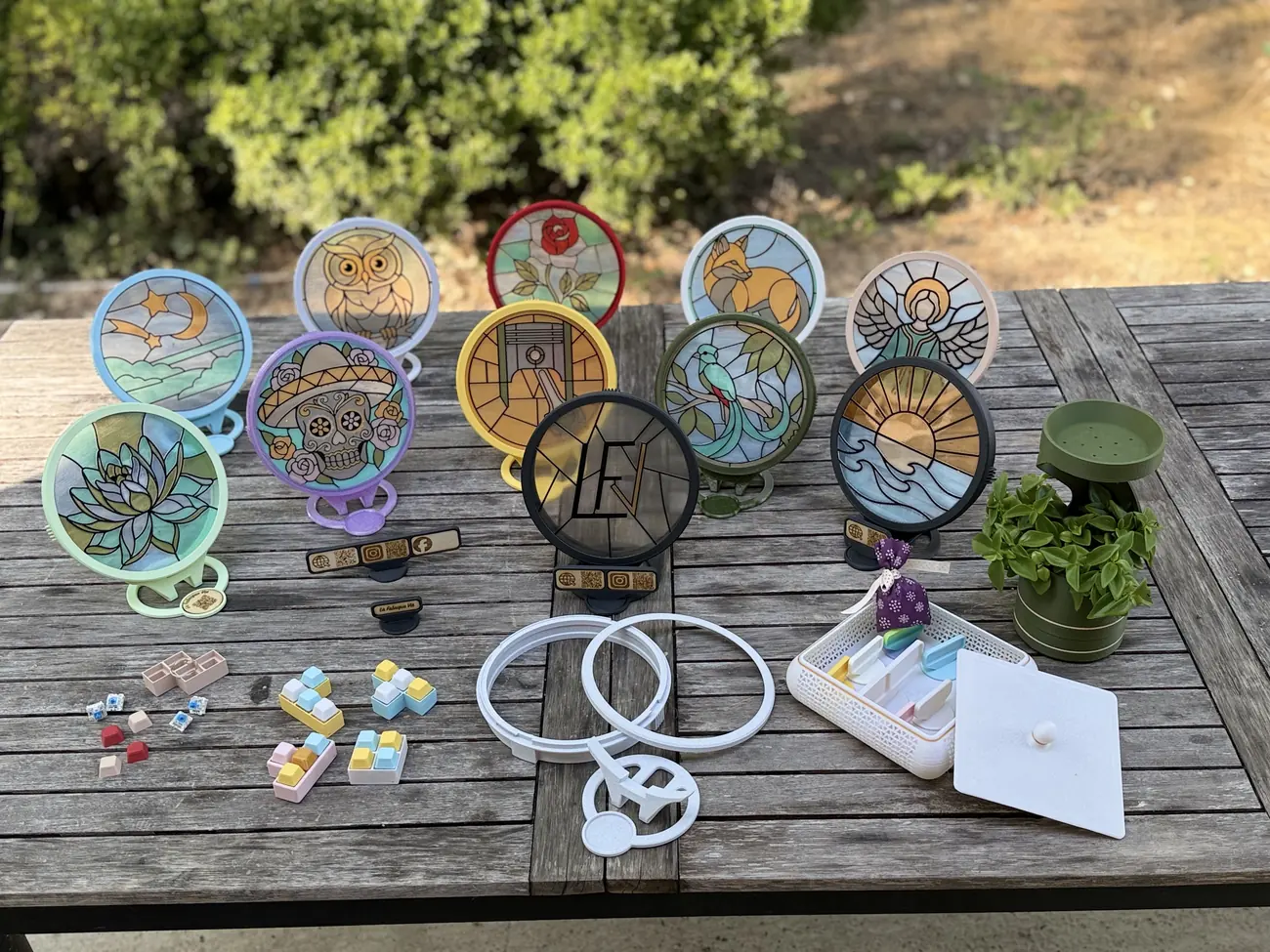 Une quinzaine de créations La Fabrique Vié posées sur une table de jardin : dix filtres Vitrail LED montés sur leurs socles — hibou, renard, rose, lotus, ange, quetzal, calavera, soleil et vagues, ciel étoilé, monogramme LFV — des plaques et porte-clés gravés, un vase spirale planté de basilic, des clickers colorés et un vide-poche.